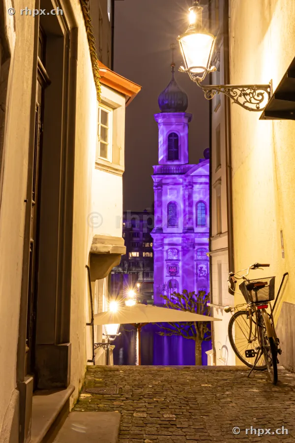 Blick durch eine Gasse auf die violett beleuchtete Jesuitenkirche während des Lilu Lichtfestivals in Luzern