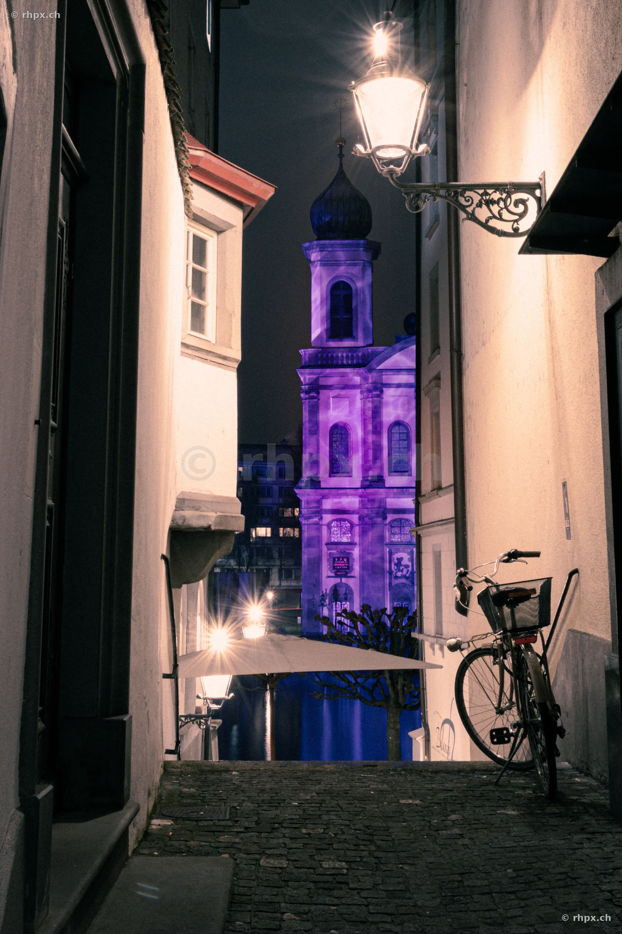 View through an alley towards the purple illuminated Jesuit Church during the Lilu Light Festival in Lucerne