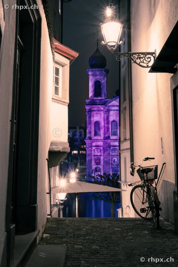 Blick durch eine Gasse auf die violett beleuchtete Jesuitenkirche während des Lilu Lichtfestivals in Luzern