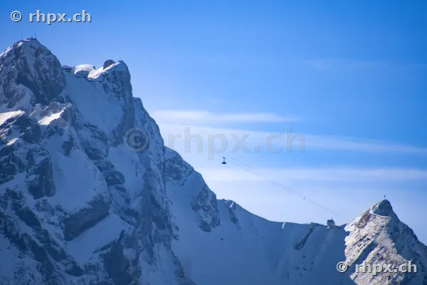 Blick auf die schneebedeckten Felswände des Pilatus mit einer Luftseilbahn vor blauem Himmel.