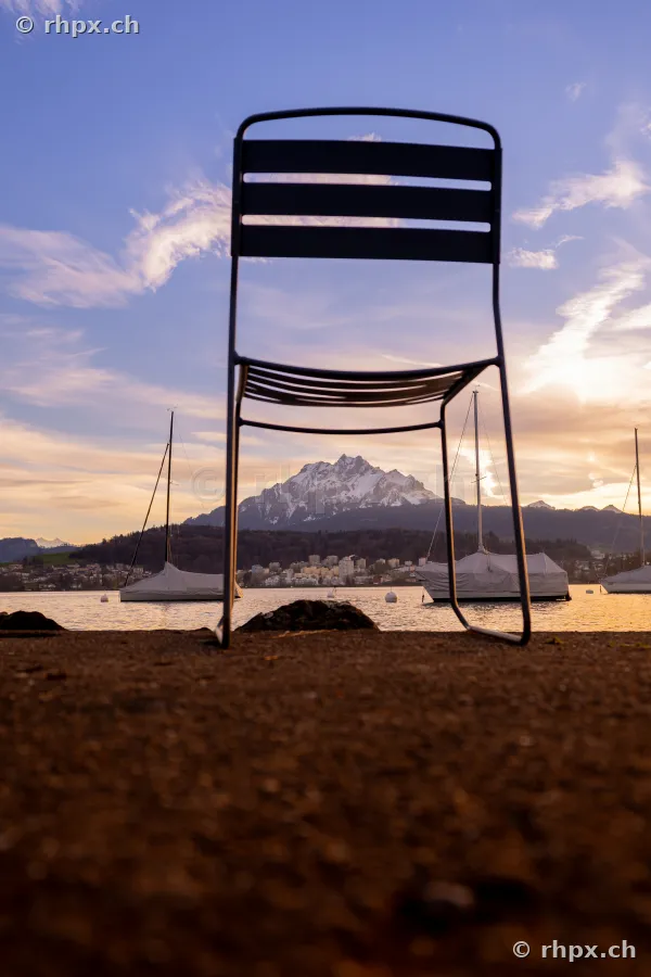 A metal chair stands on the shore in front of Lake Lucerne with a view of Mount Pilatus in the background