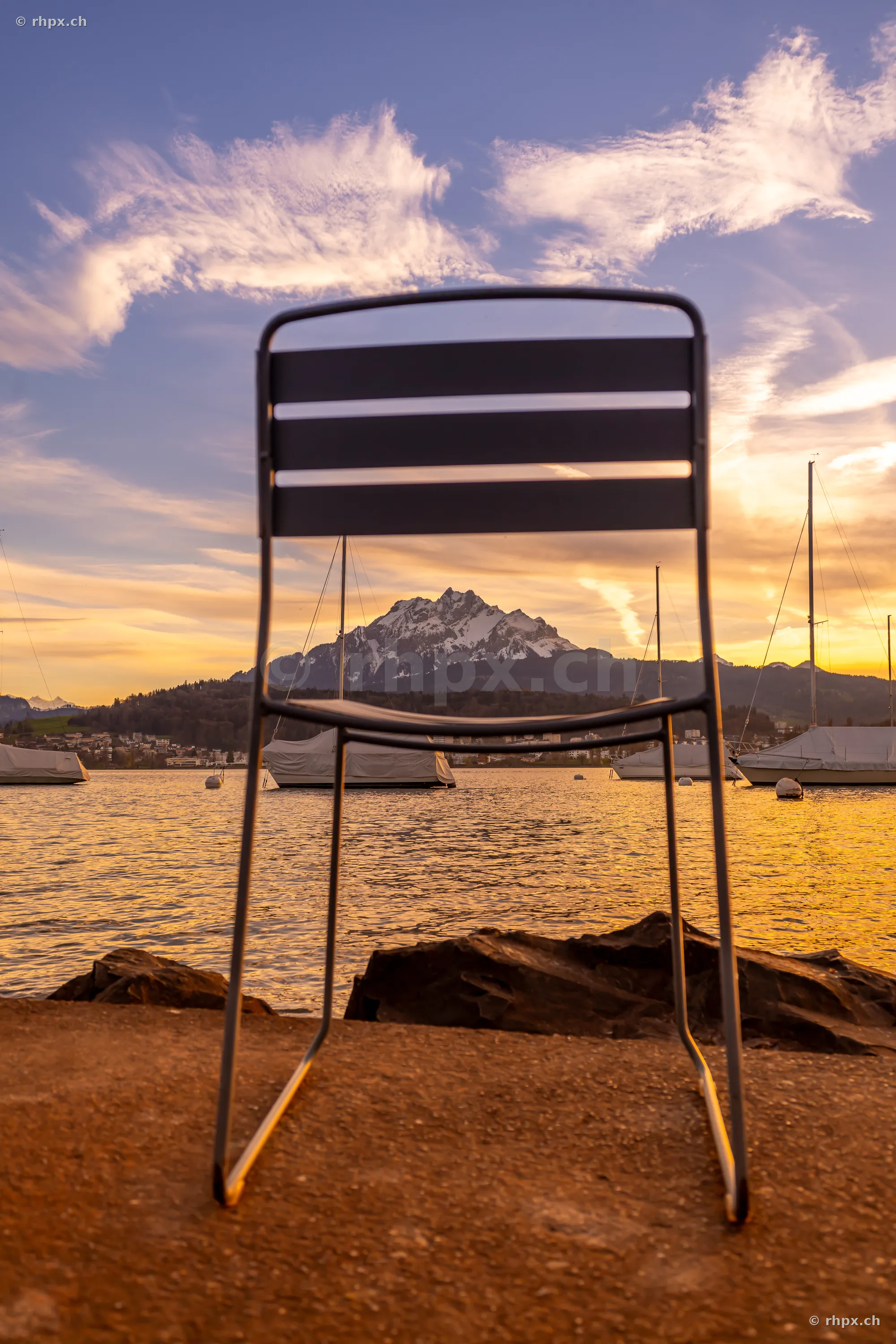 An empty chair stands on the shore in front of Lake Lucerne with Mount Pilatus in the background at sunset.