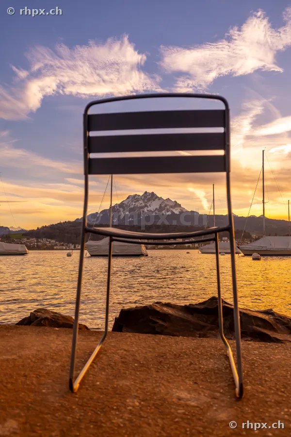 An empty chair stands on the shore in front of Lake Lucerne with Mount Pilatus in the background at sunset.