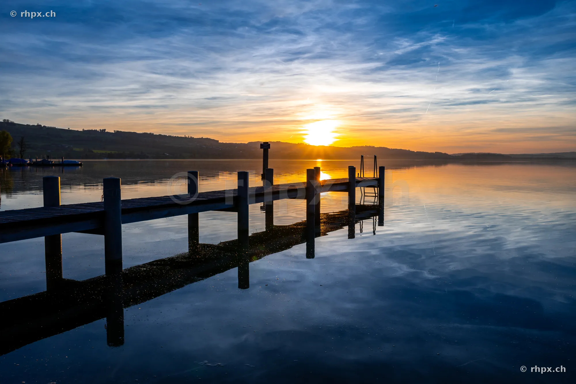 Ein langer Holzsteg führt in den ruhigen Sempachersee während eines gelb-blauen Sonnenuntergangs.