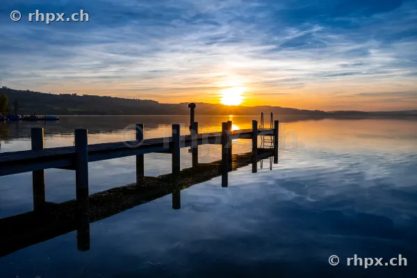 Ein langer Holzsteg führt in den ruhigen Sempachersee während eines gelb-blauen Sonnenuntergangs.