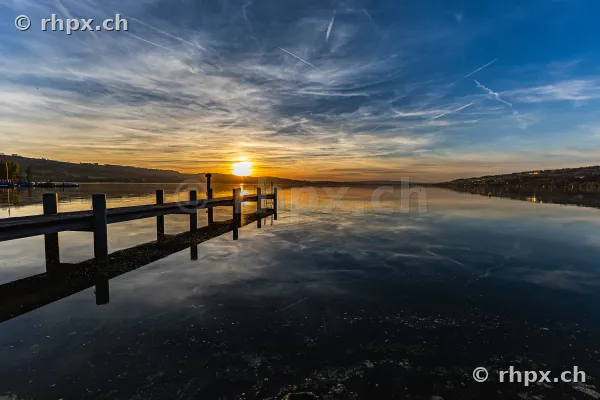 Ein hölzerner Steg führt in den ruhigen Sempachersee unter einem von Wolken durchzogenen Abendhimmel.