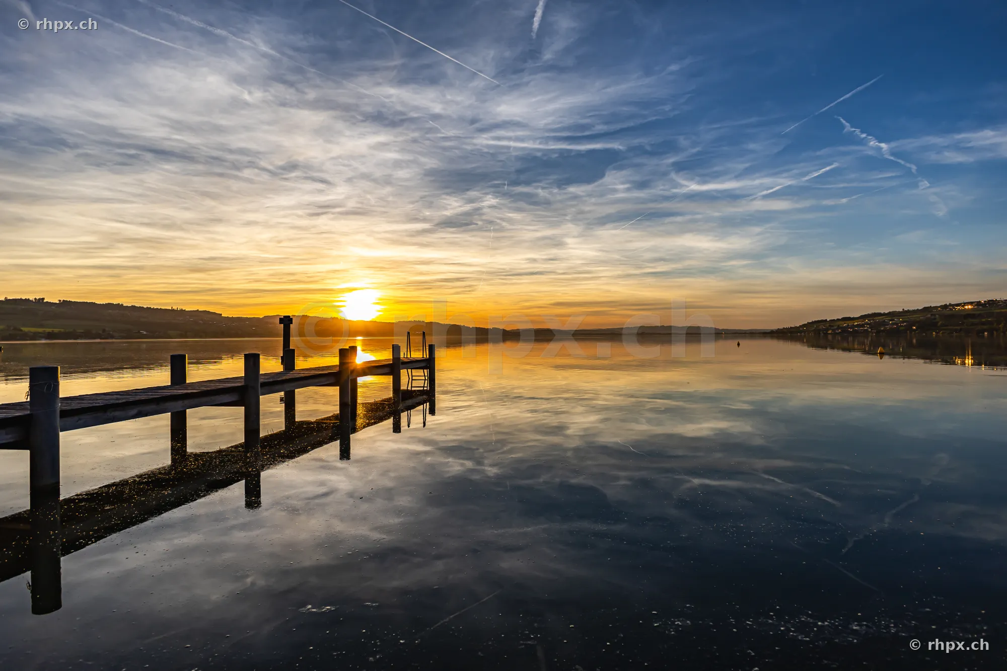 Ein hölzerner Steg führt in den ruhigen Sempachersee unter einem orangefarbenen Abendhimmel.