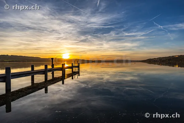 Ein hölzerner Steg führt in den ruhigen Sempachersee unter einem orangefarbenen Abendhimmel.
