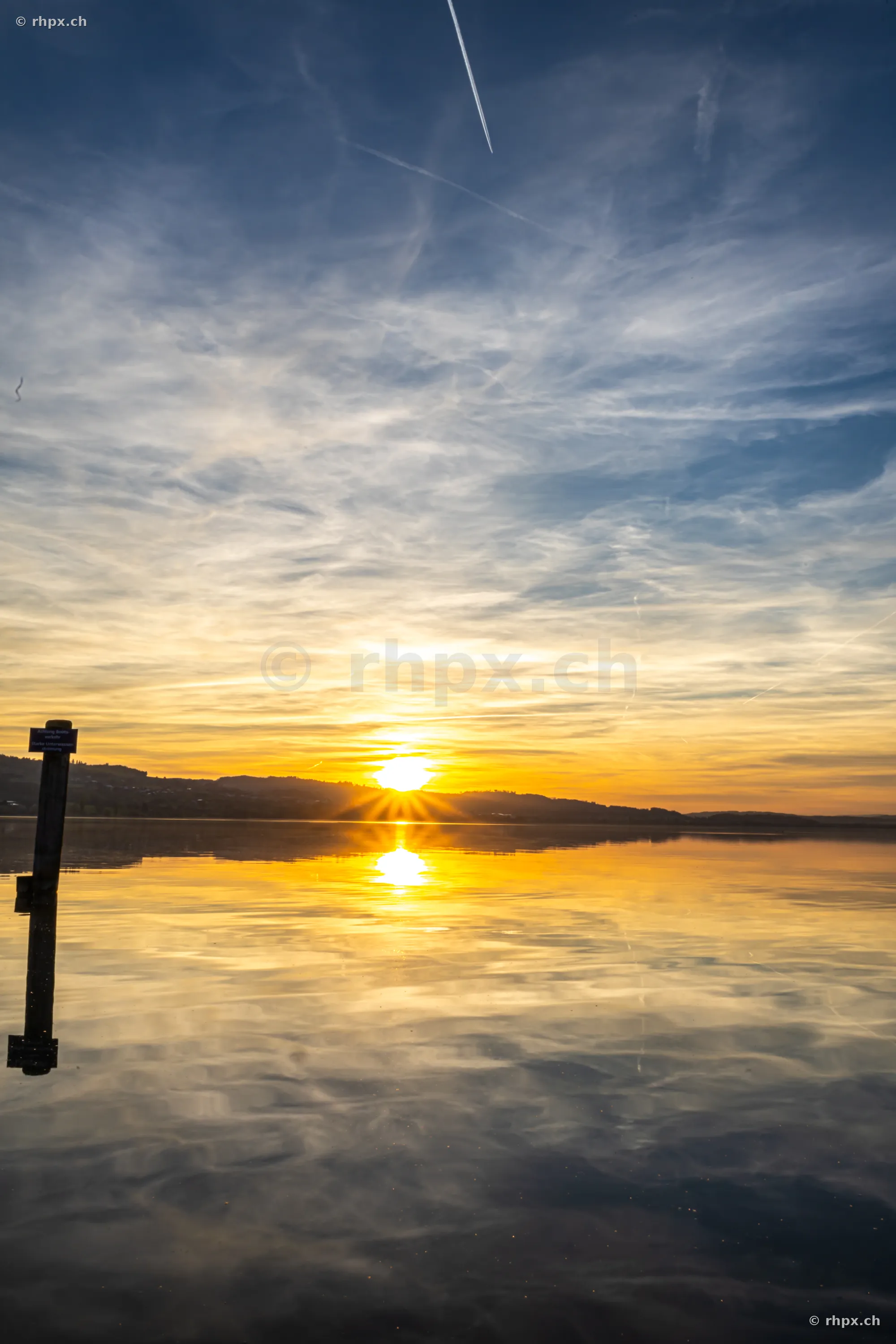 Sonnenuntergang am Sempachersee mit reflektierender Wasseroberfläche und einem Pfahl im Vordergrund