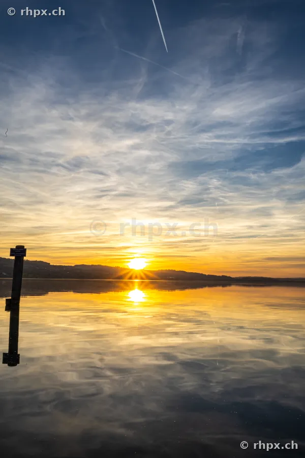 Sonnenuntergang am Sempachersee mit reflektierender Wasseroberfläche und einem Pfahl im Vordergrund