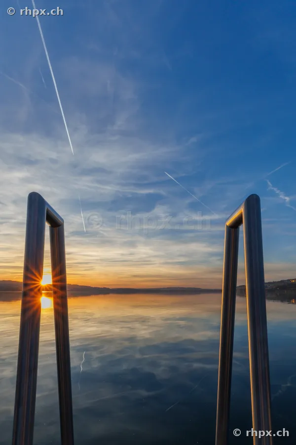 Blick durch eine metallene Badeleiter über den ruhigen Sempachersee beim Sonnenuntergang