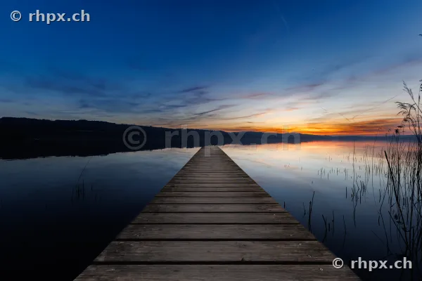 Ein gerader Holzsteg führt in die ruhige Wasseroberfläche des Sempachersees bei Sonnenuntergang