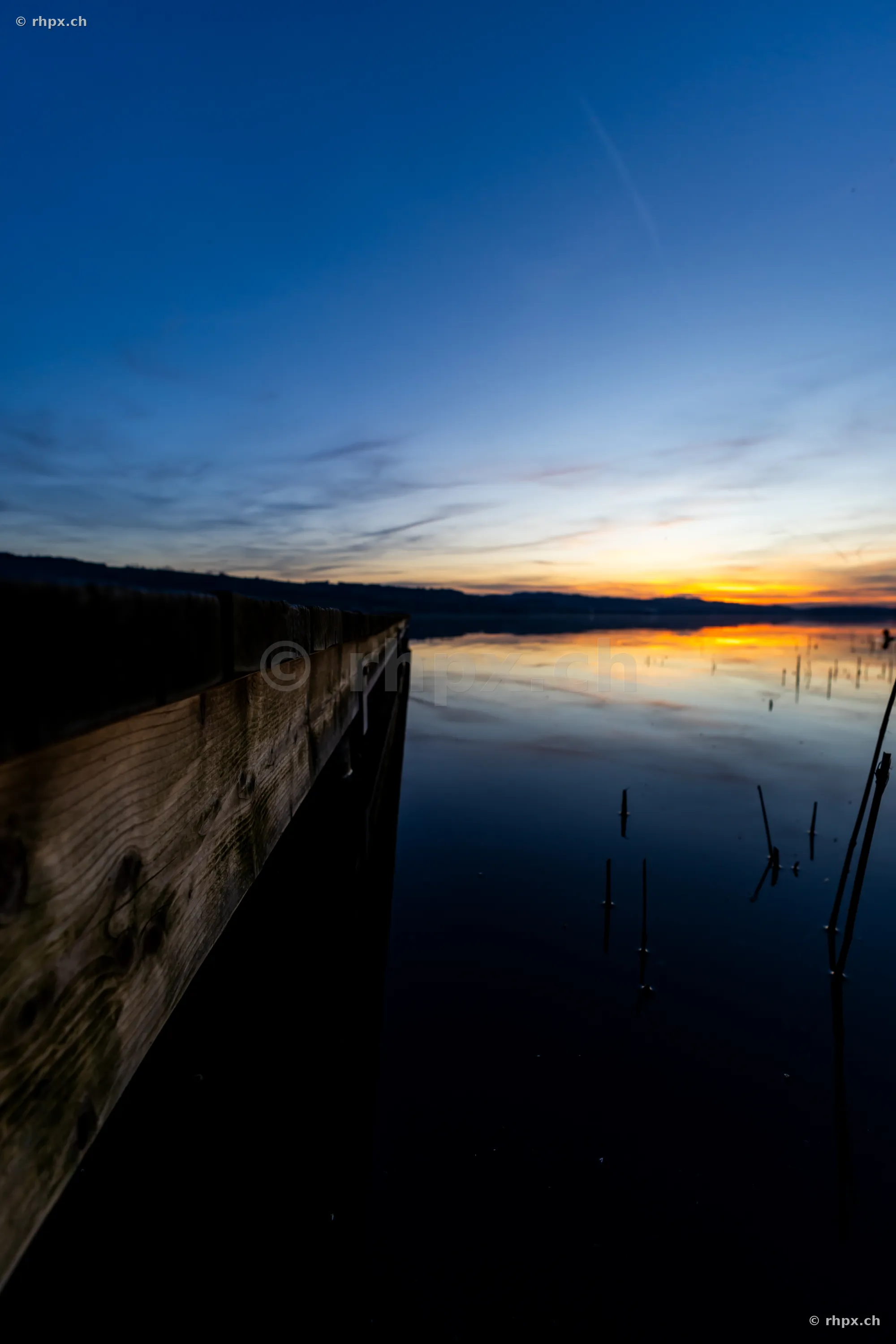 Ein hölzerner Bootssteg führt in das ruhige Wasser des Sempachersees bei einsetzender Abenddämmerung.