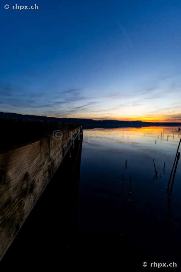 Ein hölzerner Bootssteg führt in das ruhige Wasser des Sempachersees bei einsetzender Abenddämmerung.