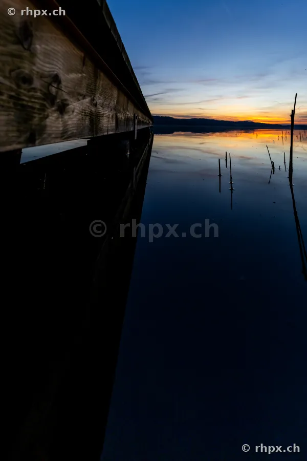 Blick entlang eines hölzernen Bootsstegs auf den ruhigen Sempachersee während des Sonnenuntergangs