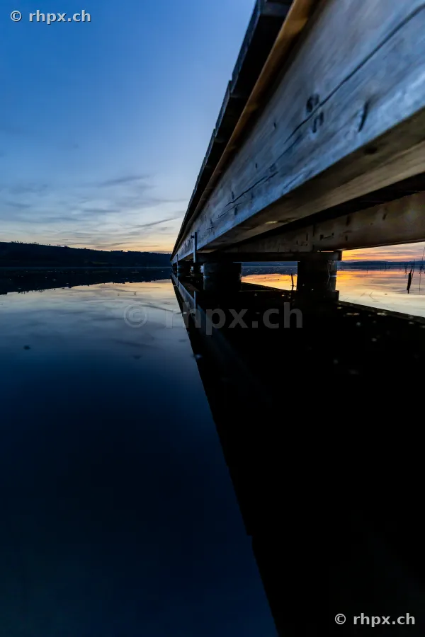 Blick unter einem Holzsteg am Sempachersee mit Spiegelung im Wasser bei Dämmerung