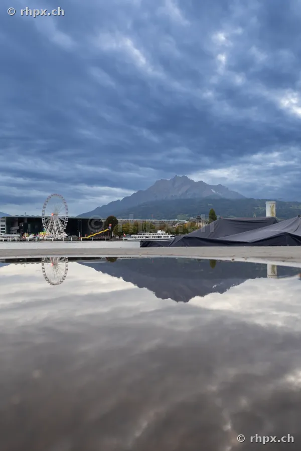 Riesenrad und Berg Pilatus spiegeln sich in einer Pfütze am Ufer des Vierwaldstättersees in Luzern.