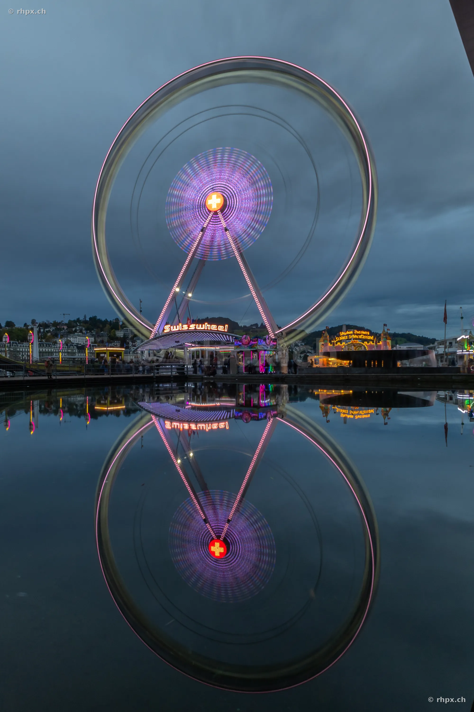 The Swisswheel Ferris wheel at twilight on the lakeshore in Lucerne, reflected in the water surface.