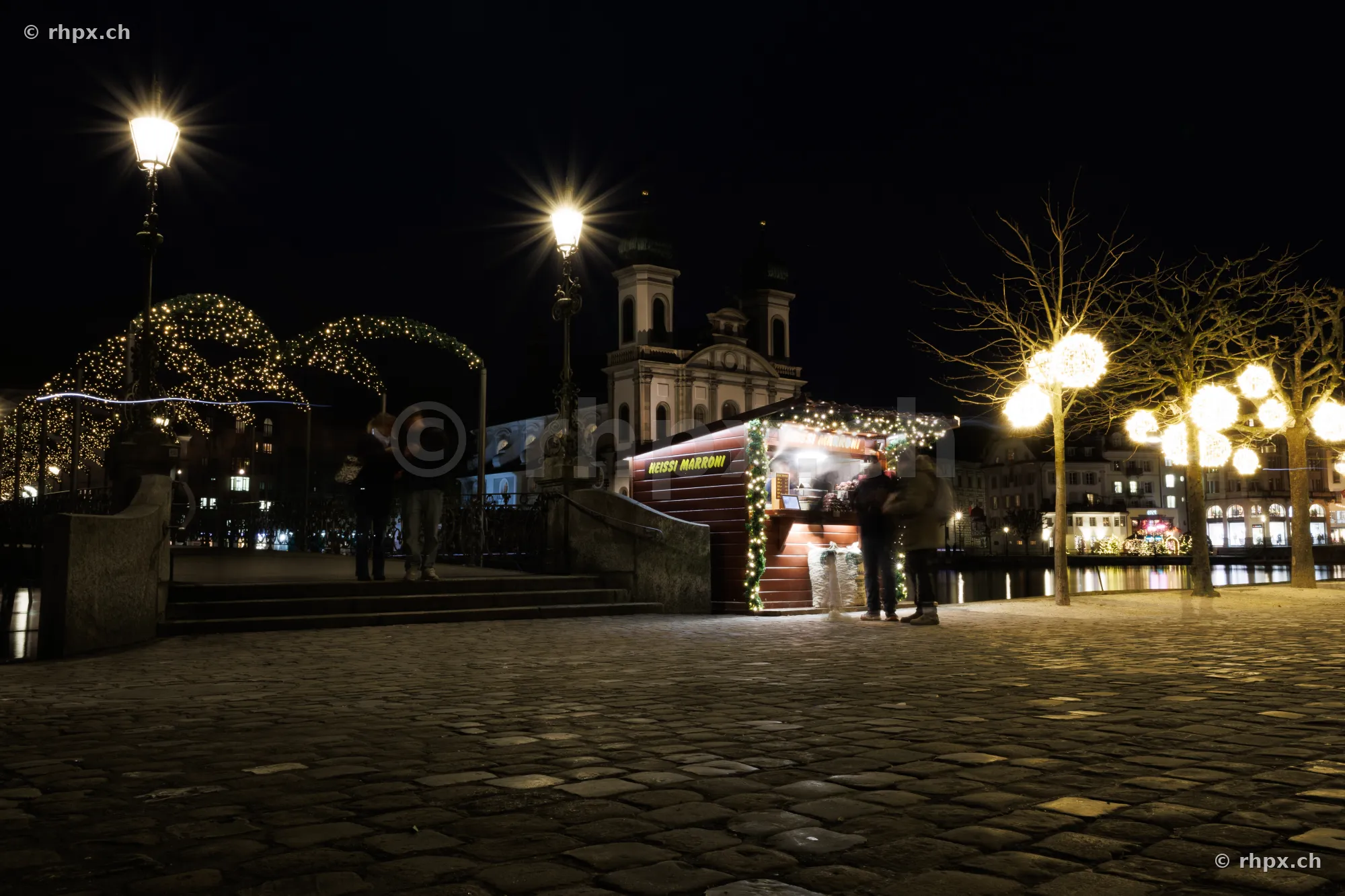 Weihnachtsbeleuchtung und Marronistand am Ufer der Reuss in Luzern bei Nacht mit der Jesuitenkirche im Hintergrund