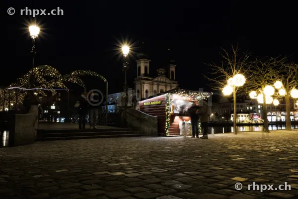Weihnachtsbeleuchtung und Marronistand am Ufer der Reuss in Luzern bei Nacht mit der Jesuitenkirche im Hintergrund