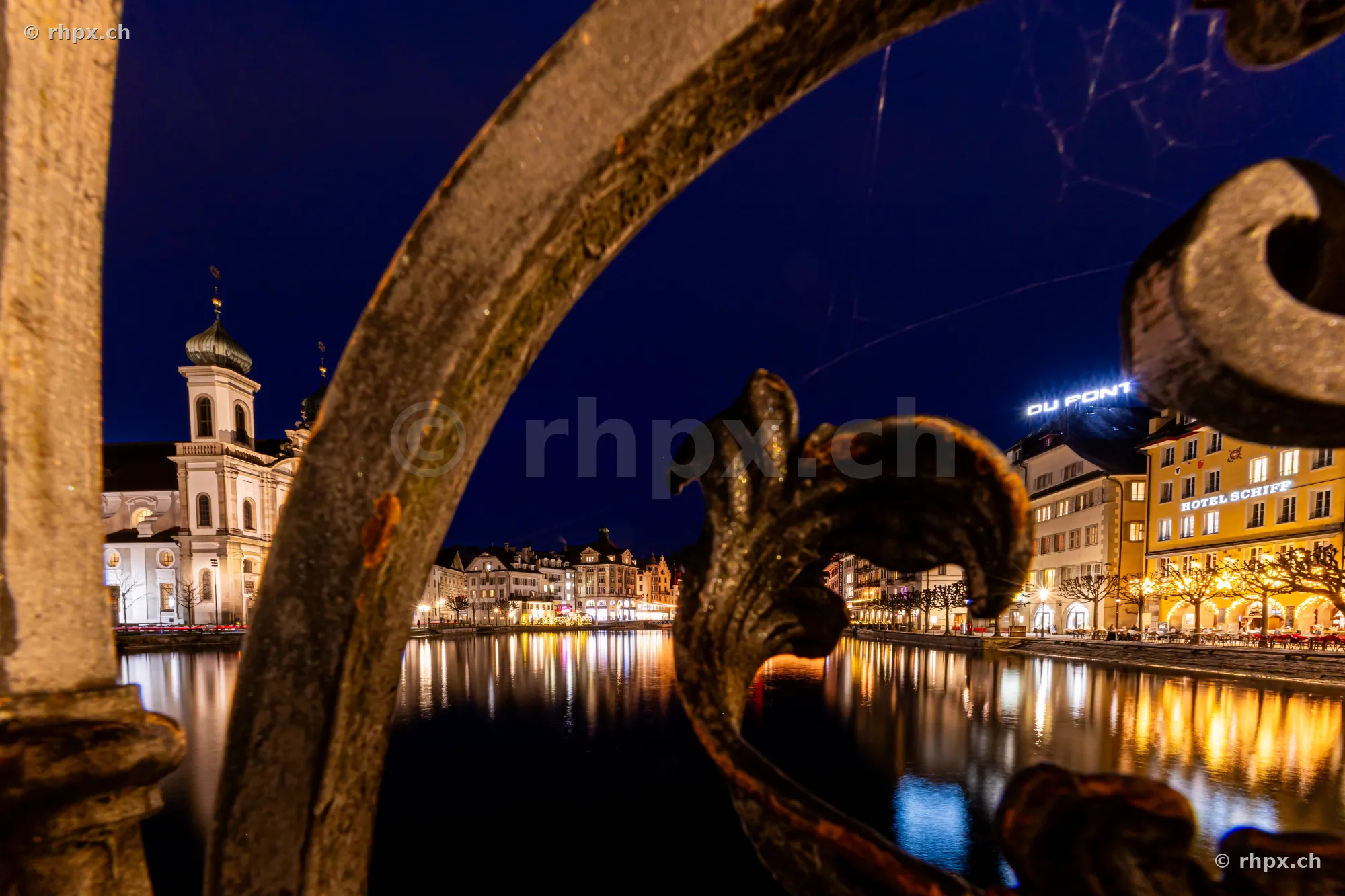 Blick durch ein verziertes Eisengeländer auf die nächtliche Jesuitenkirche und die Uferpromenade in Luzern
