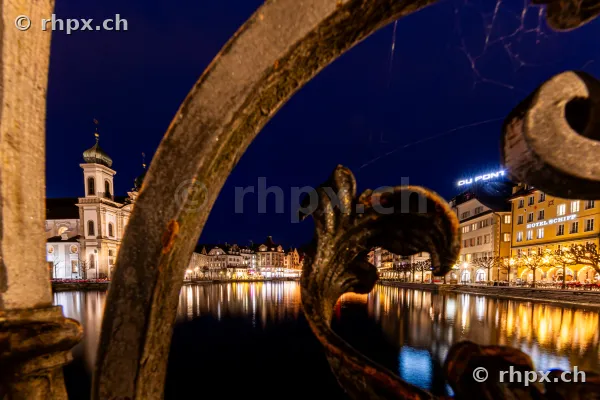 View through a decorative iron railing towards the illuminated Jesuit Church and riverbank in Lucerne at night