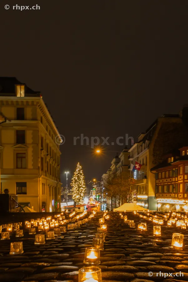 Many small candles on dark cobblestones in front of a night cityscape in Lucerne.