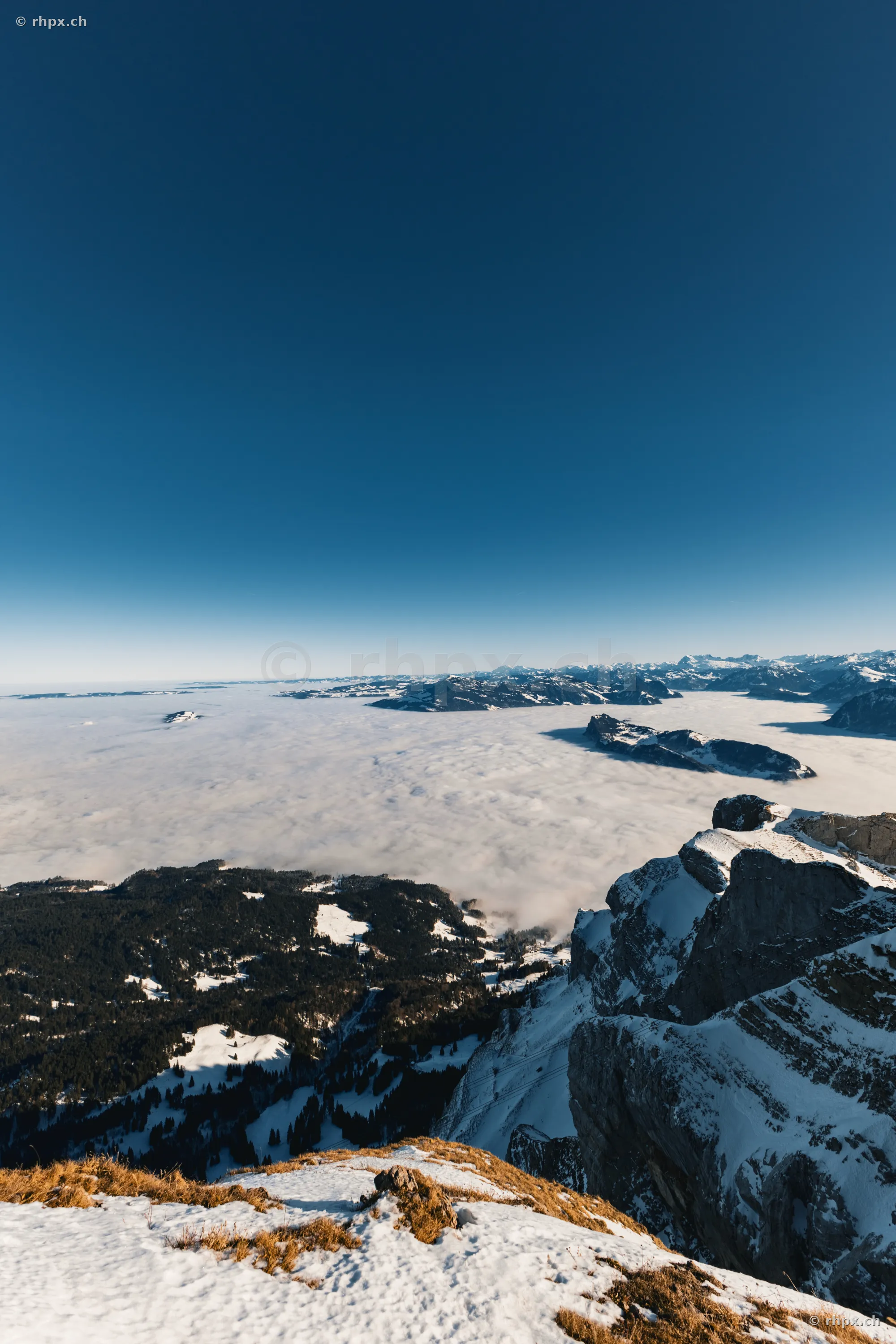 Blick von der verschneiten Felsflanke des Pilatus über ein tief unterhalb liegendes Nebelmeer bei klarem Himmel.