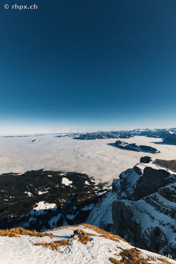 Blick von der verschneiten Felsflanke des Pilatus über ein tief unterhalb liegendes Nebelmeer bei klarem Himmel.