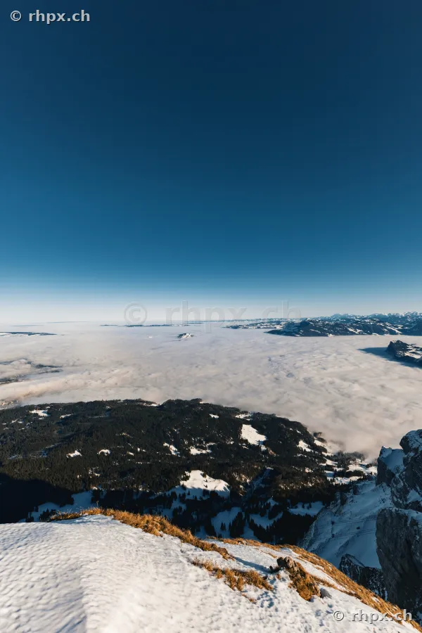 Blick vom schneebedeckten Gipfel des Pilatus auf ein unterhalb liegendes Wolkenmeer bei klarem Himmel.