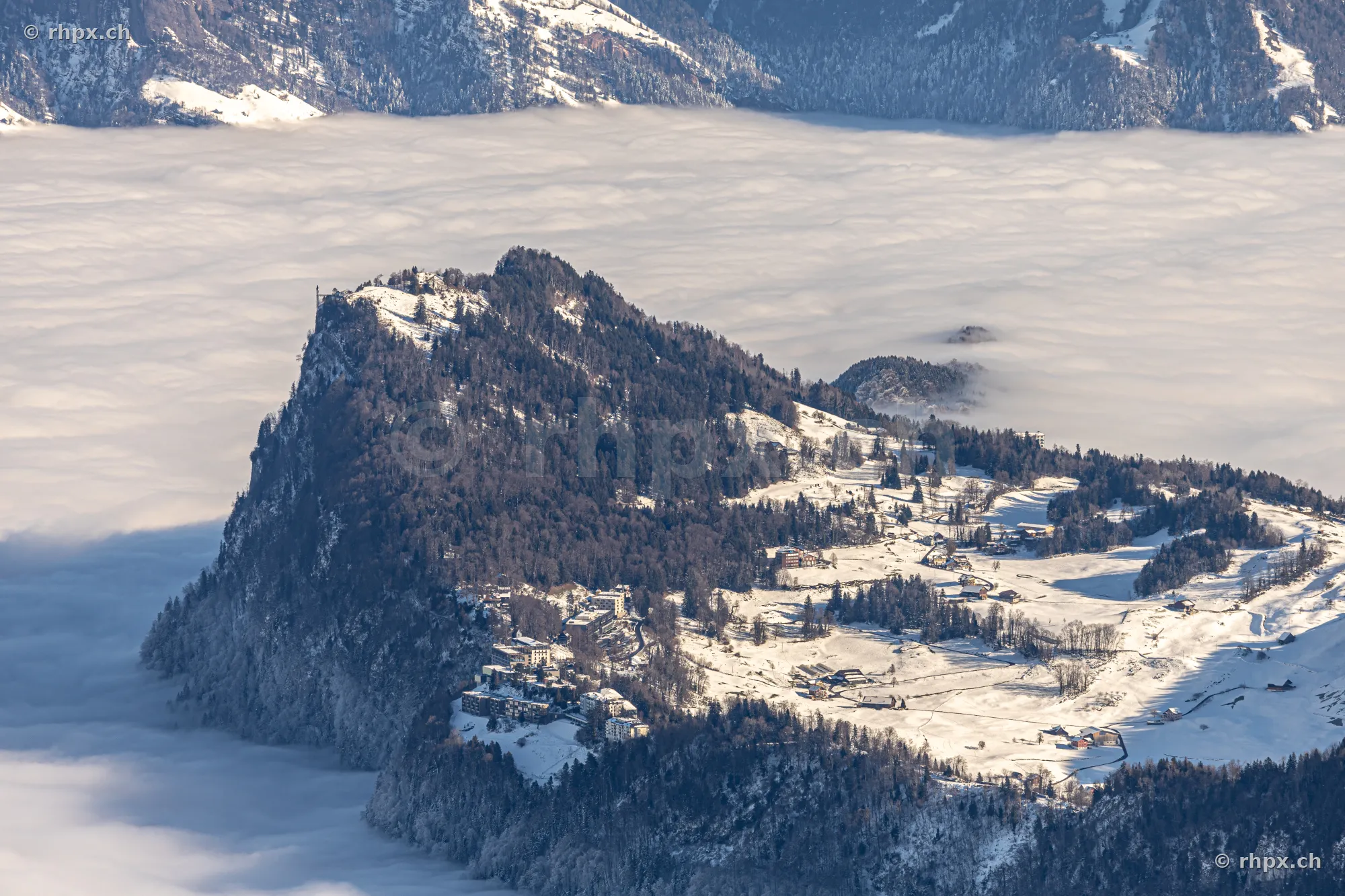 Luftaufnahme des verschneiten Bürgenstocks mit Hotellerie über einer tief liegenden Nebeldecke.