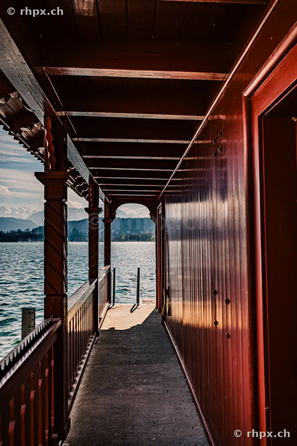 View along a covered wooden pier towards Lake Lucerne with the Alps in the background.