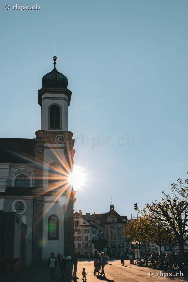 The Jesuit Church in Lucerne illuminated by the low sun with pedestrians walking on the square in front.