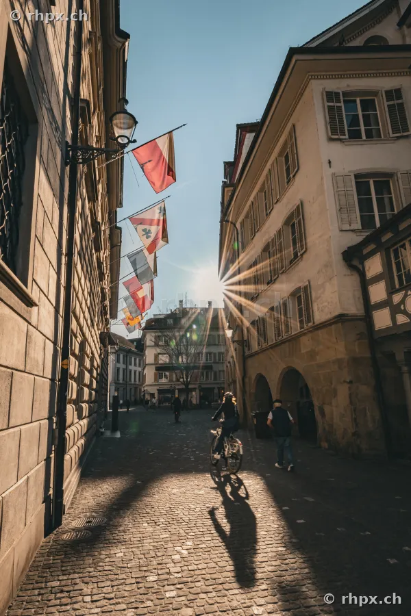 People and a cyclist in a narrow alley in Lucerne Old Town with backlighting.