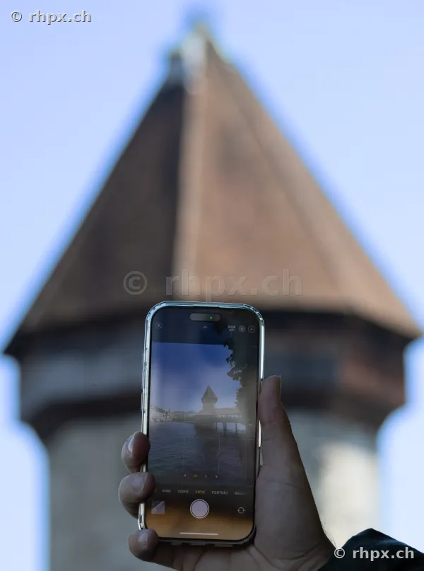 Hand hält ein Smartphone, das den Wasserturm der Kapellbrücke in Luzern fotografiert, vor dem unscharfen Hintergrund des realen Turms.