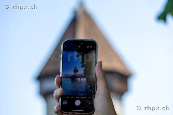 A hand holding a smartphone displaying the Chapel Bridge in Lucerne, with the blurred water tower in the background.