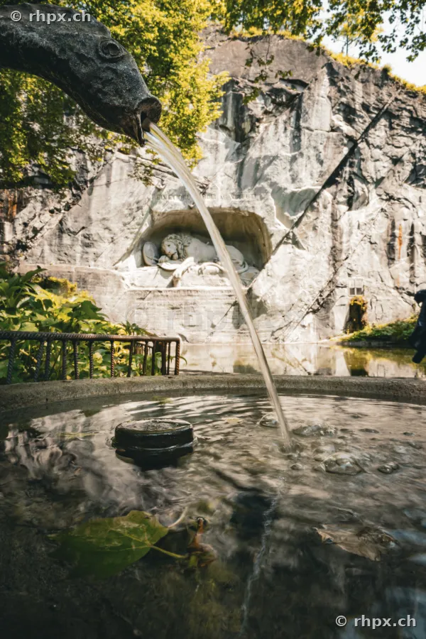 Fountain with fish figure in the foreground and the Lion Monument in Lucerne in the background