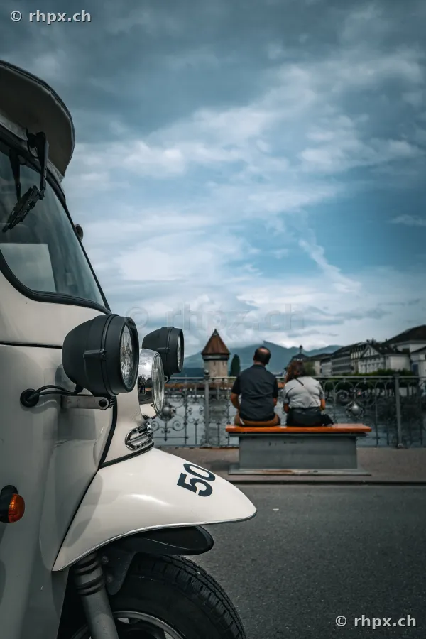 Detailansicht eines dreirädrigen Ape-Fahrzeugs mit einem Paar, das auf einer Parkbank vor der Kapellbrücke in Luzern sitzt.