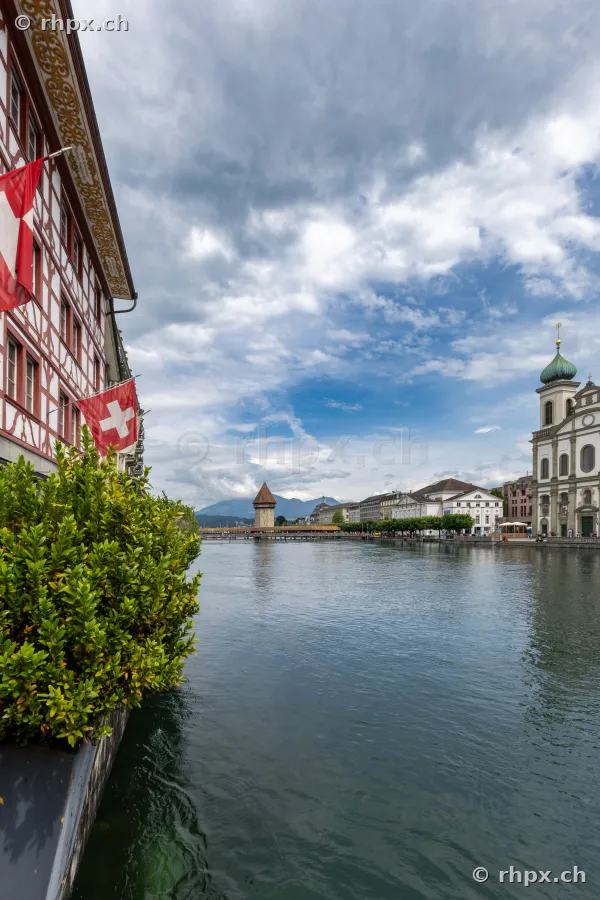 View of the Reuss river in Lucerne with Chapel Bridge, Jesuit Church and Swiss flags on the left bank