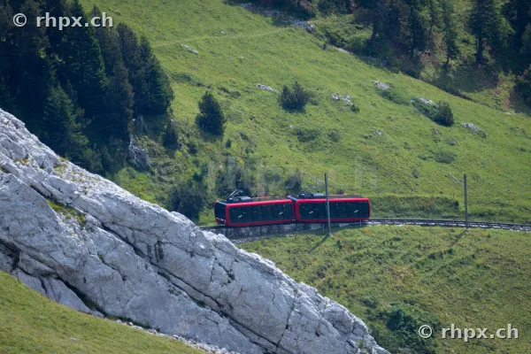 Roter Triebwagen der Pilatusbahn fährt an einer steilen Felswand entlang über grüne Bergwiesen