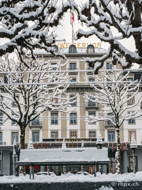 Vorderansicht des Hotel Schweizerhof Luzern im Winter mit verschneiter Parkbank im Vordergrund