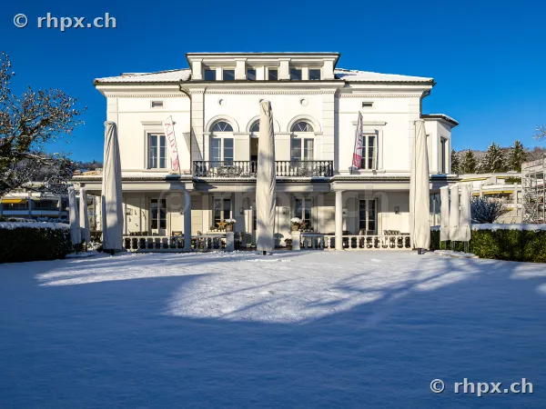 Die weisse Villa Schweizerhof in Luzern vor einer schneebedeckten Fläche unter blauem Himmel.