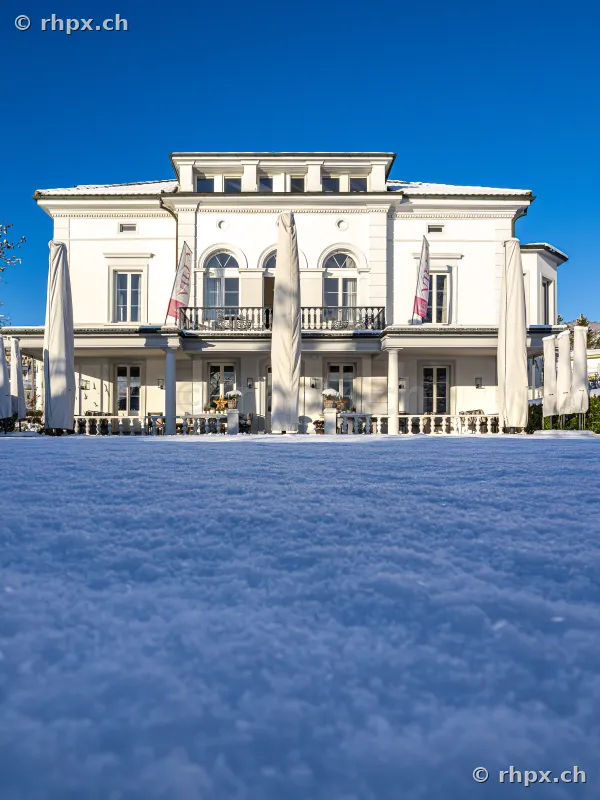 Weisse Fassade der Villa Schweizerhof in Luzern vor einer schneebedeckten Fläche unter blauem Himmel.