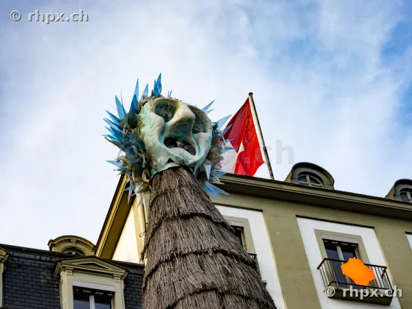 Eine grosse Fasnachtsmaske mit strohbedecktem Körper vor der Fassade des Hotel Schweizerhof in Luzern mit Schweizer Fahne