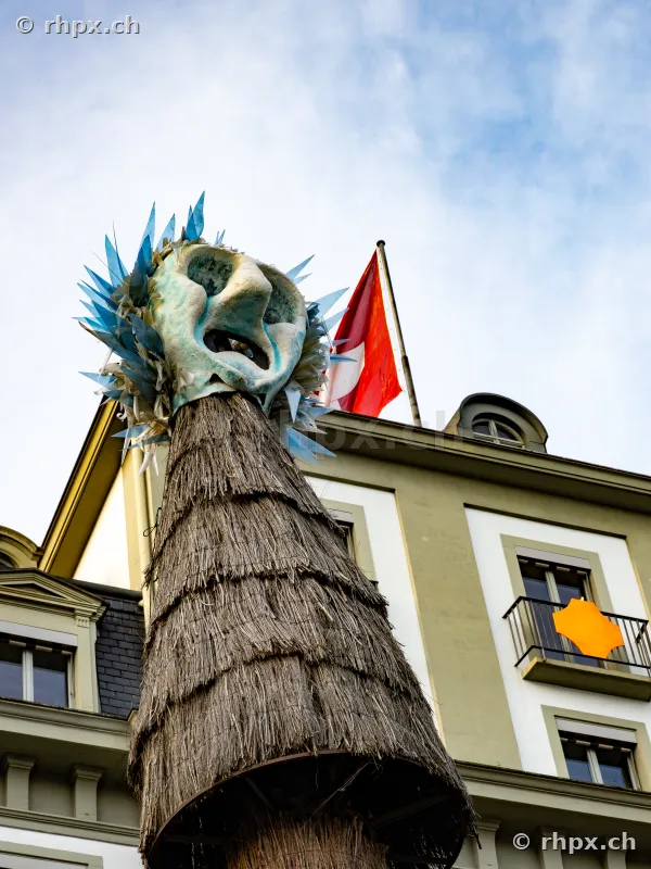 Eine grosse Fasnachtsfigur mit Strohkörper vor dem Hotel Schweizerhof in Luzern mit Schweizer Fahne im Hintergrund.