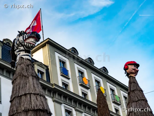Zwei grosse Fasnachtsfiguren mit Strohkörpern vor der Fassade des Hotel Schweizerhof in Luzern unter blauem Himmel.