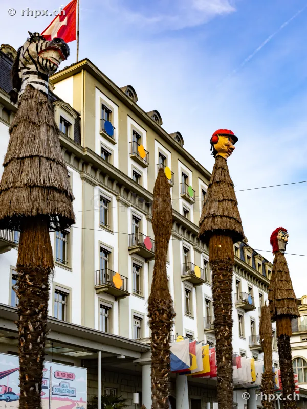 Mit Stroh verkleidete Palmen und Fasnachtsfiguren vor dem Hotel Schweizerhof in Luzern unter blauem Himmel