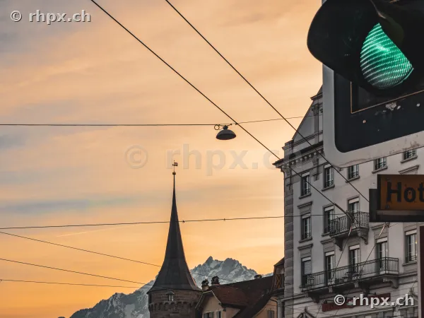 Green traffic light, with a tower of Lucerne's old town and snowy Alps in the background at sunset.