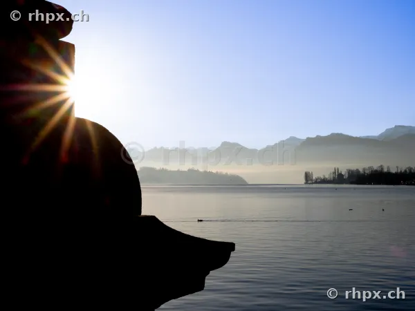 Sonneneinstrahlung hinter einem dunklen Steinelement am Vierwaldstättersee mit Blick auf die Alpen