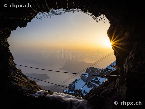 View through a rock opening at Pilatus overlooking the snowy Alps and Lake Lucerne at sunset